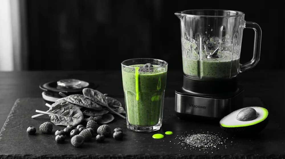 Grüner Smoothie mit Spinat, Beeren, Chiasamen und Avocado neben einem Hochleistungsmixer auf heller Küchenarbeitsfläche
